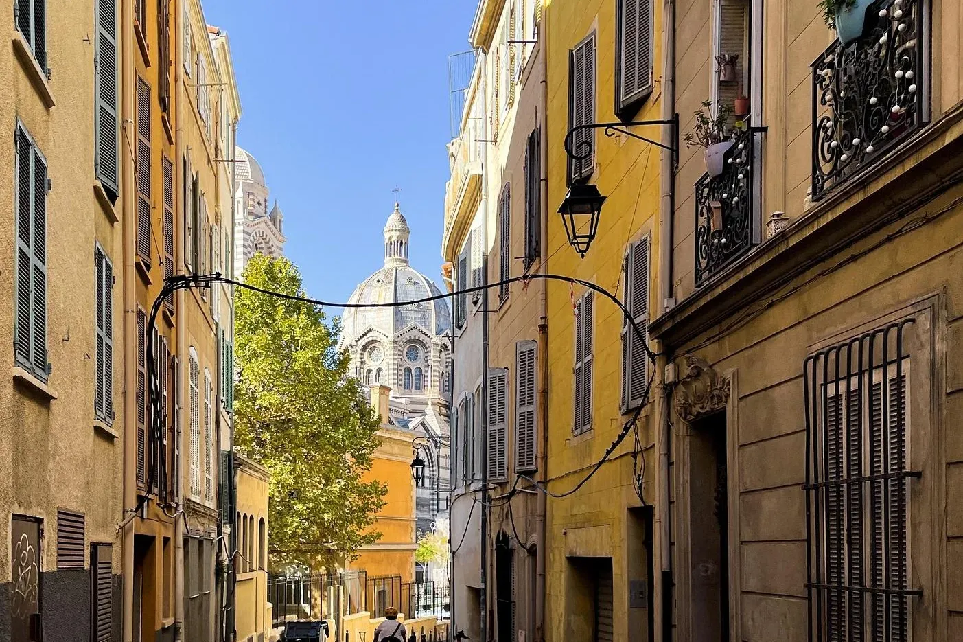 Narrow street in Marseille lined with tall yellow buildings with shutters, leading towards the dome of Notre-Dame de la Ga...