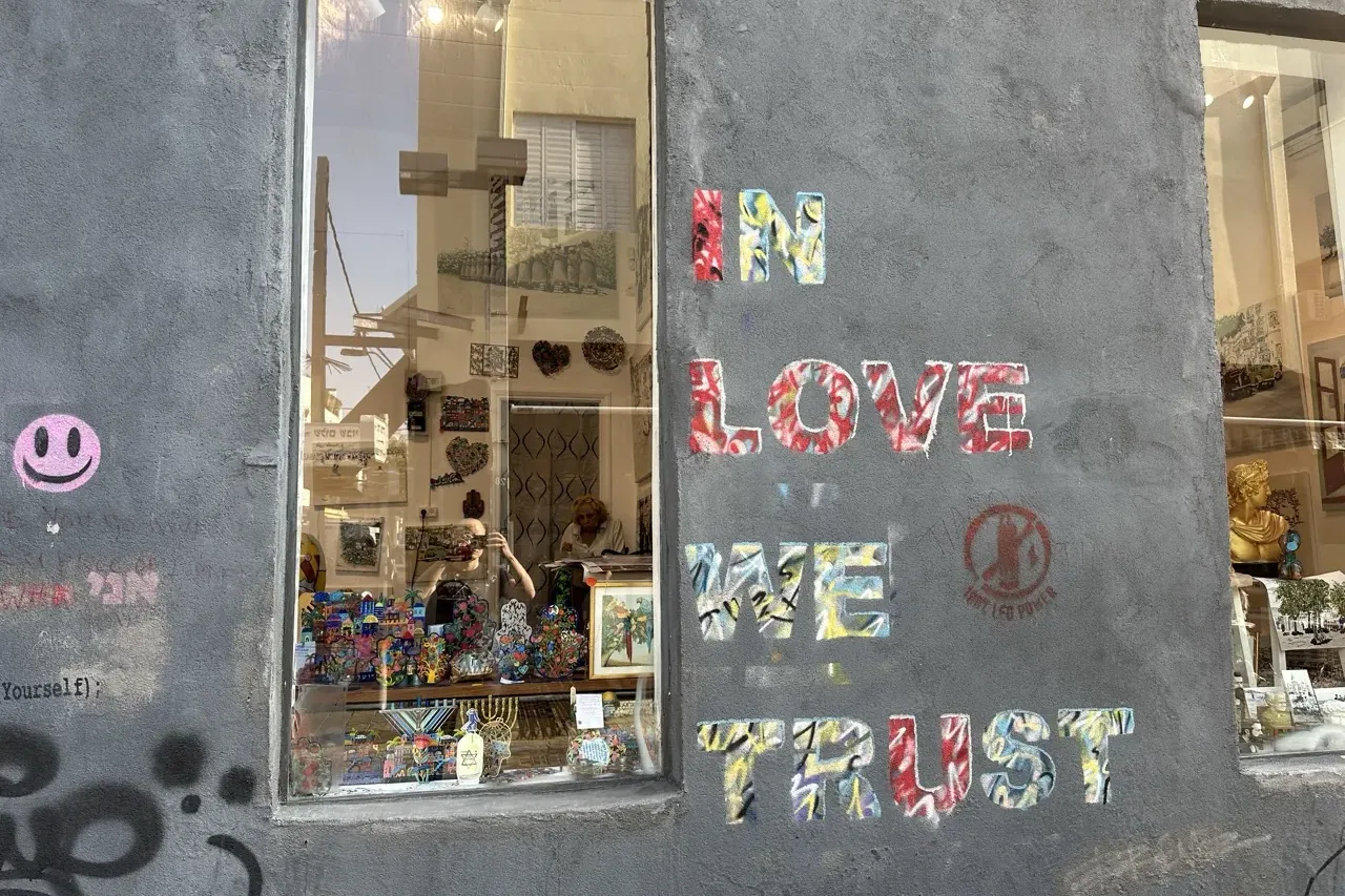Grey wall with colorful "IN LOVE WE TRUST" graffiti, a pink smiley face, and a shop window reflecting buildings.