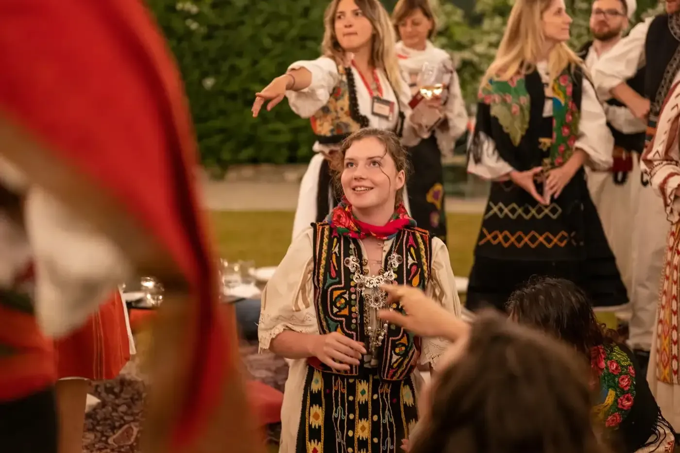 Young woman in embroidered traditional Albanian clothing and a red scarf smiling, with other people in traditional attire ...