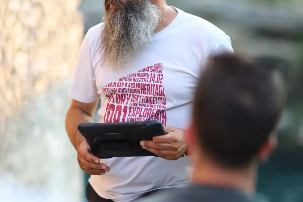 A bearded male tour guide with sunglasses on his head holds a tablet, speaking to a group outdoors in Mostar.