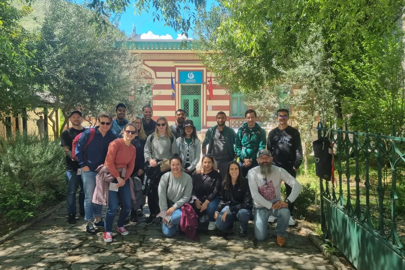 Group of 16 tourists posing at the Yunus Emre Institute in Mostar, a red and white striped building surrounded by trees.