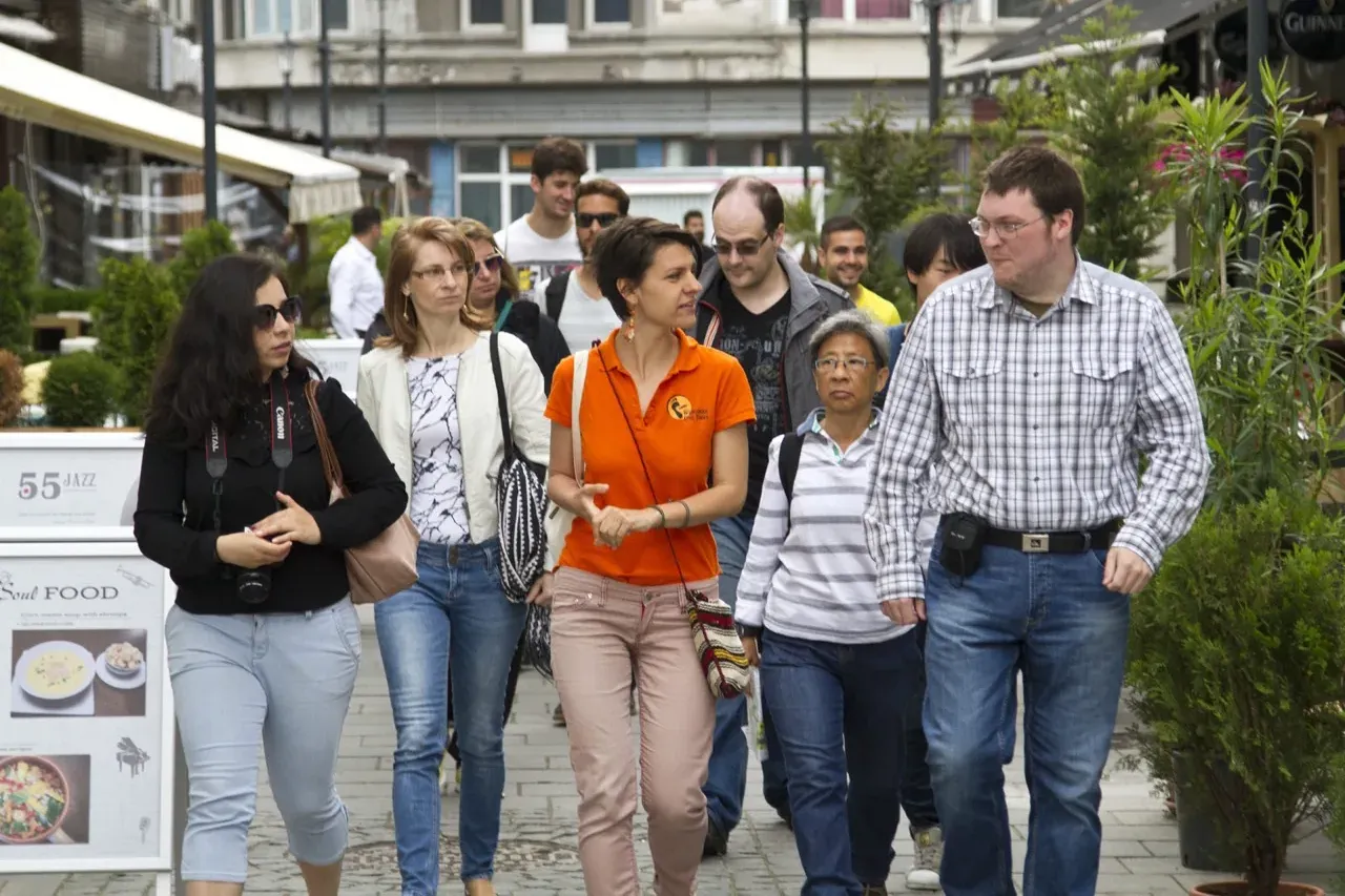 A diverse group of people, including a guide in an orange polo shirt, walking on a paved street in Bucharest, past buildin...