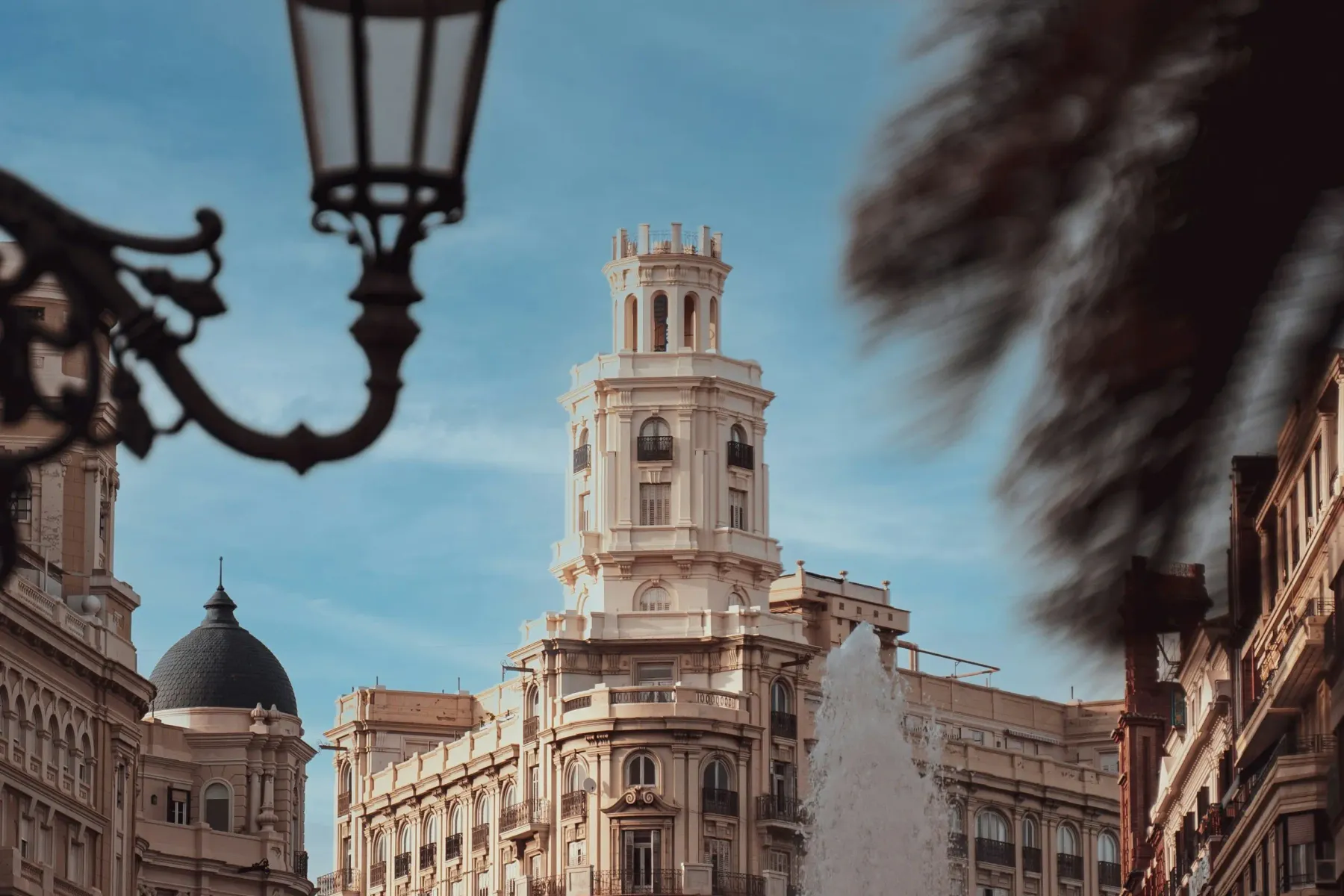 Historic buildings in Valencia, including a prominent tower, seen from a street. A large fountain sprays water, with ornat...