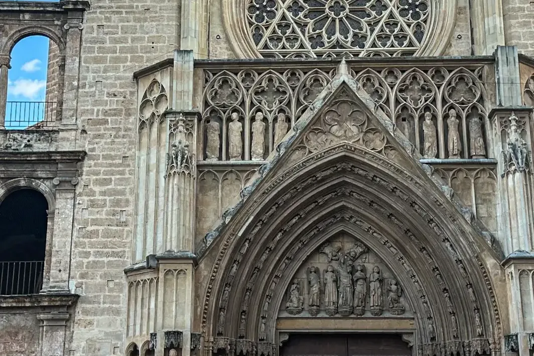 Gothic stone facade of Valencia Cathedral with a large rose window, ornate arched portal, and sculpted figures under blue ...