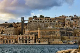 View of Valletta's historic fortifications and stone buildings along the Grand Harbour, with a dramatic cloudy sky above.