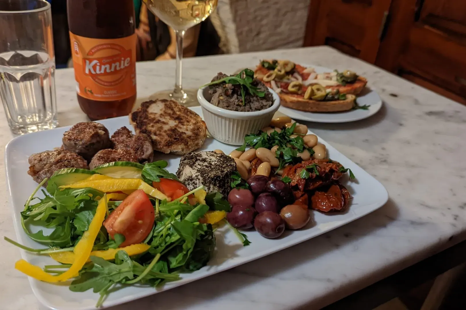 A platter of Maltese food including meatballs, salad, beans, olives, and cheese, with a Kinnie bottle and wine glass on a ...