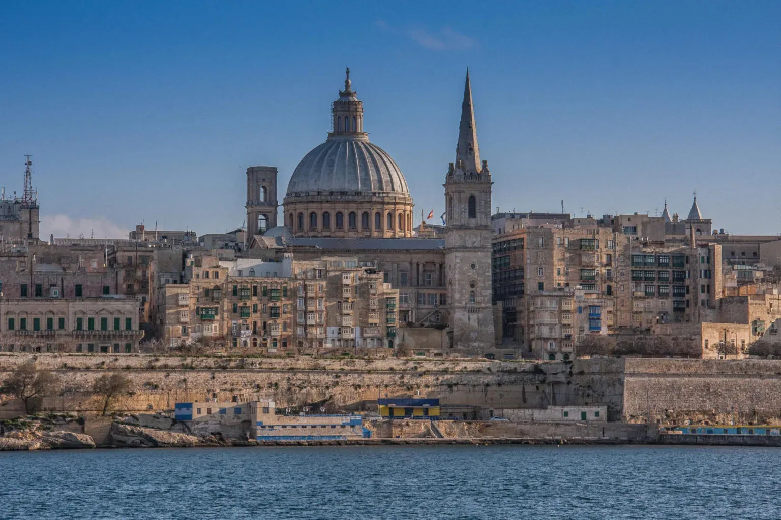 Valletta cityscape from the water, featuring a large dome, a tall spire, and historic buildings along a rocky shore.