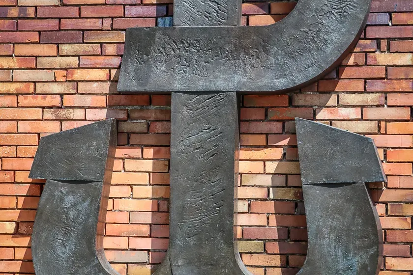 Large, dark metal sculpture of the Fighting Poland (Kotwica) symbol on a multi-colored brick wall with green leaves above ...