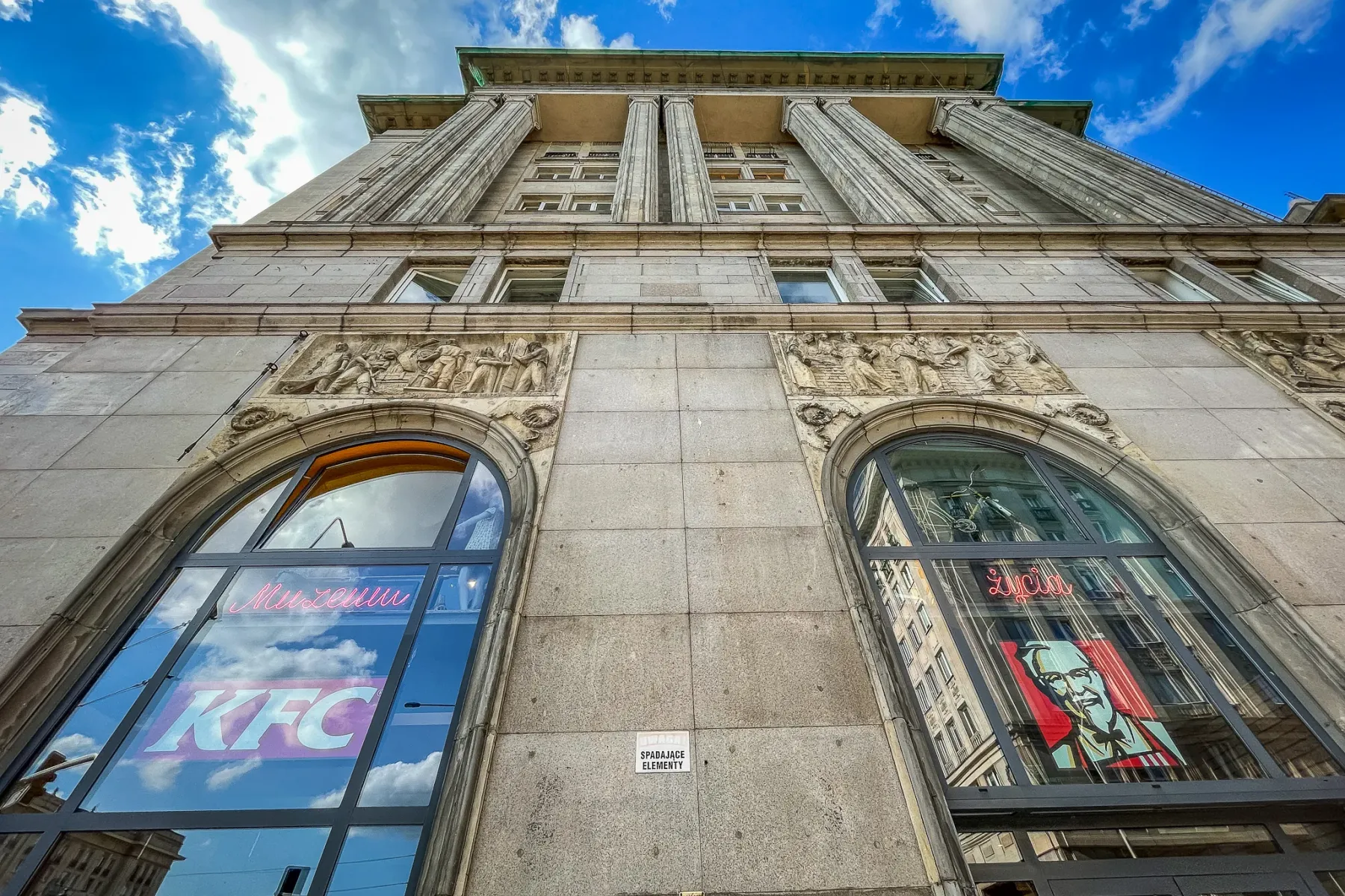 Low-angle view of a grey stone building with classical columns and relief carvings. Large arched windows show reflections,...