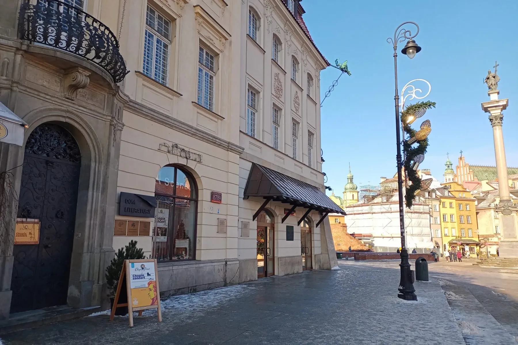 Facade of Dom Literatury (House of Literature) and buildings in Warsaw's Old Town, with a Chopin concert sign. Snowy pavem...