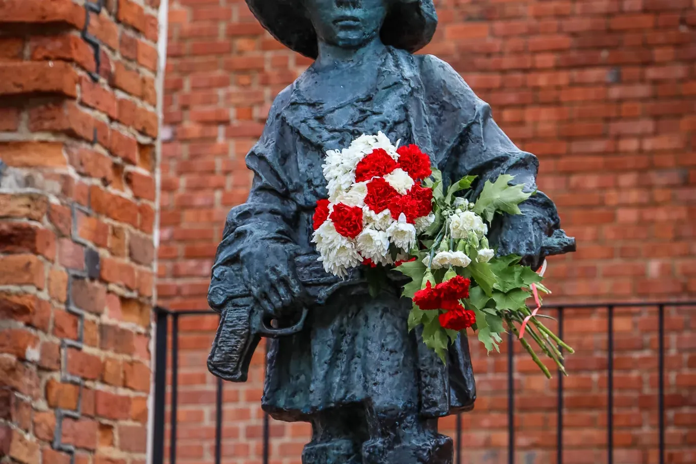 Bronze statue of a child soldier in a helmet holding red and white flowers, against a brick wall.