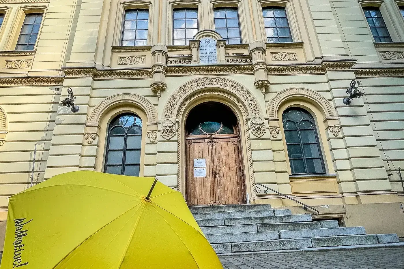 Front facade of the ornate Nożyk Synagogue in Warsaw, a yellow Walkative umbrella on stone steps in the foreground.