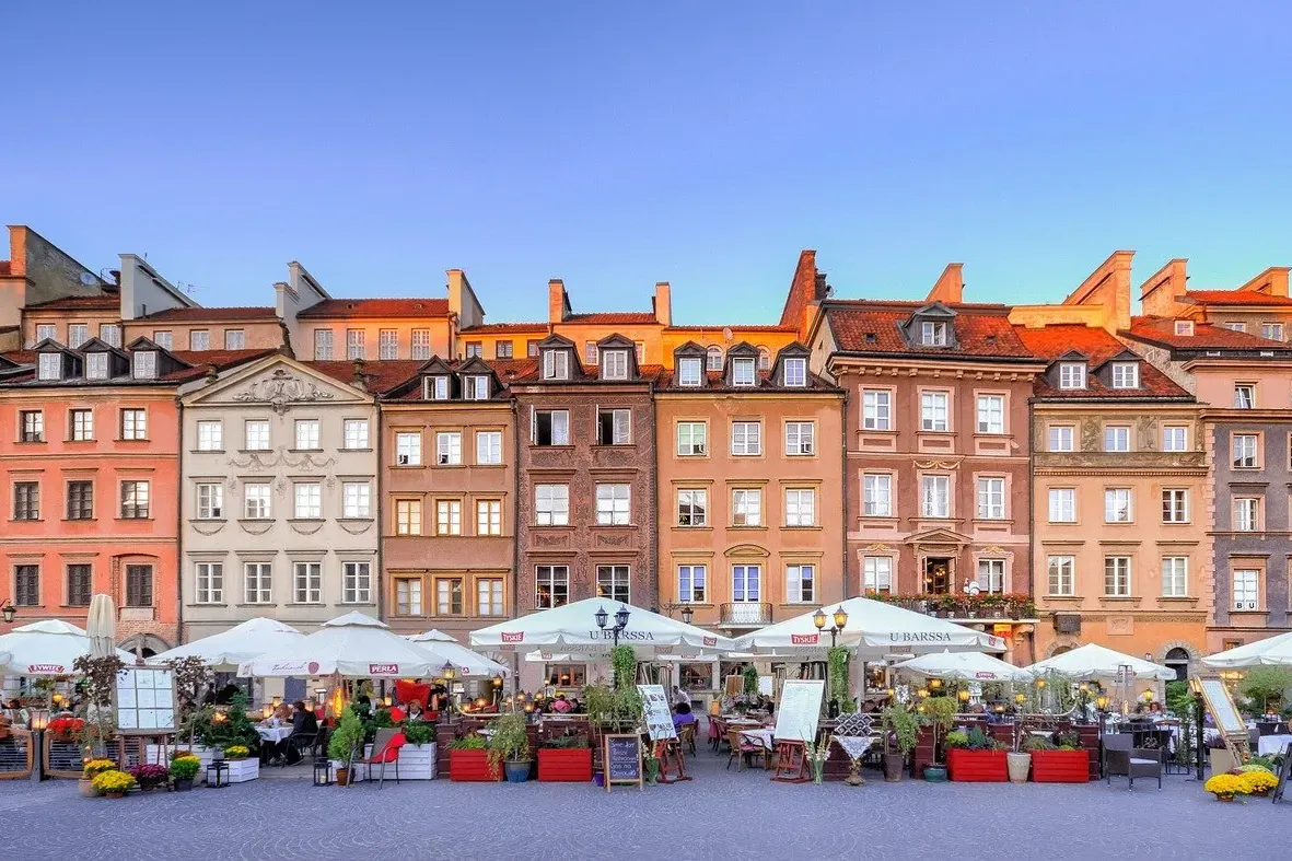 Colorful historic buildings with red roofs overlook an outdoor cafe area with white umbrellas and people dining under a bl...