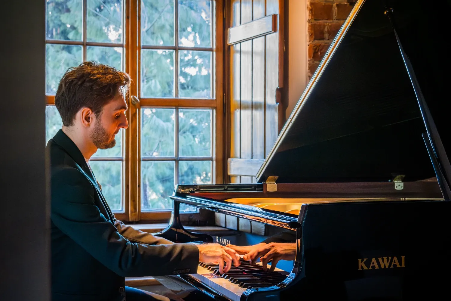 Man in a suit playing a black Kawai grand piano next to a window with wooden shutters.