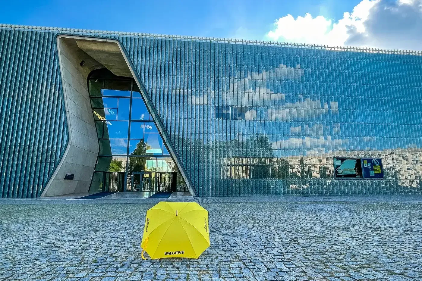 Yellow Walkative umbrella on a cobblestone plaza in front of the modern glass exterior of POLIN Museum in Warsaw under a b...