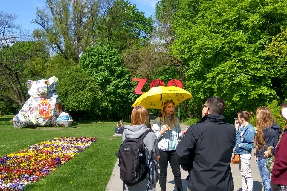 Tour guide with yellow umbrella speaking to a group on a paved path, next to a flower bed and green trees. A ZOO sign and ...