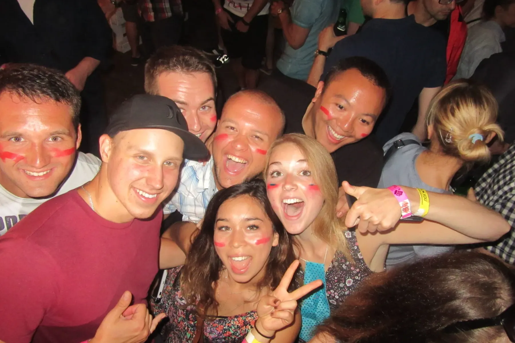 A diverse group of young people smiling, many with red face paint on their cheeks, posing closely together at a lively ind...