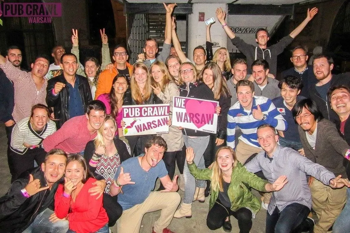 A large group of diverse young adults smiling, posing, and holding "Pub Crawl Warsaw" and "We love Warsaw" signs in an urb...