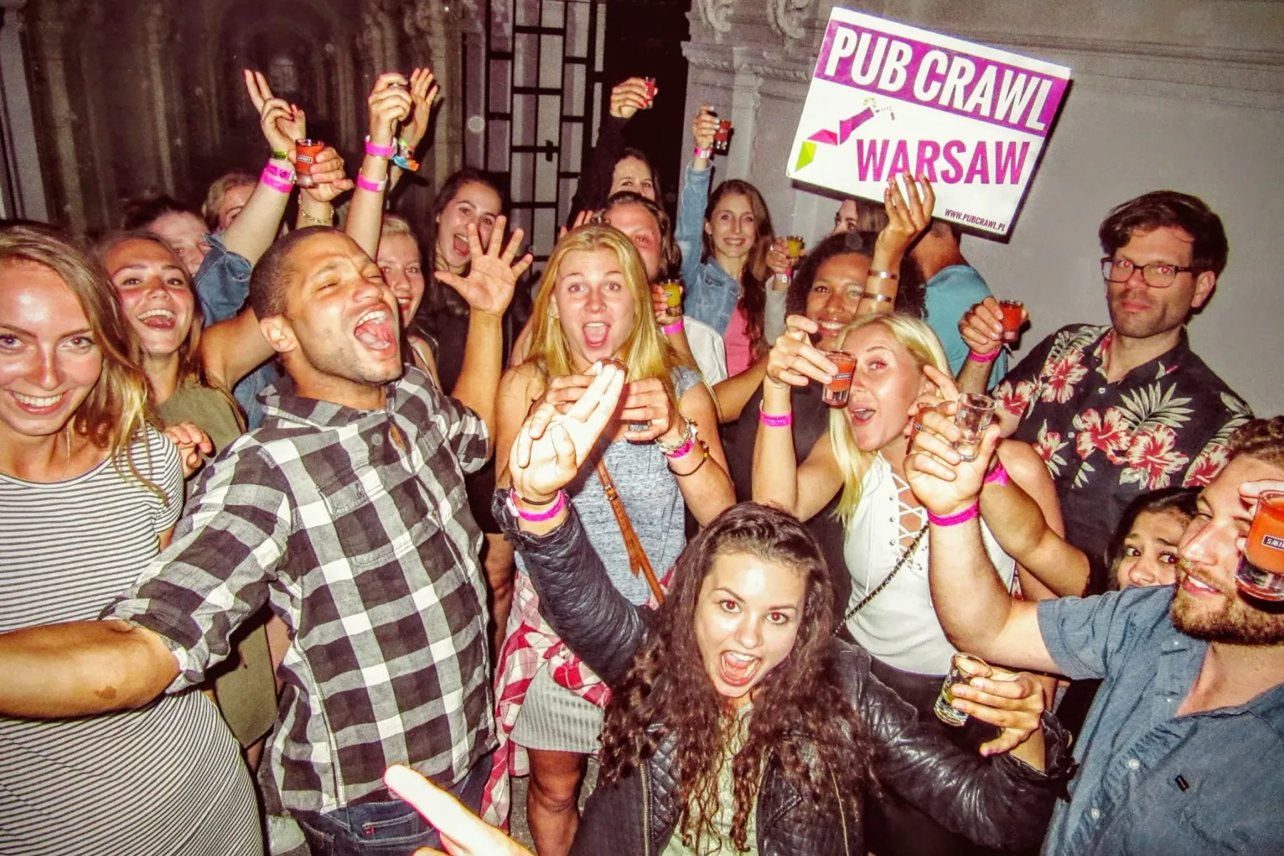 Group of young adults smiling, laughing, and holding shot glasses, one holding a 'PUB CRAWL WARSAW' sign.