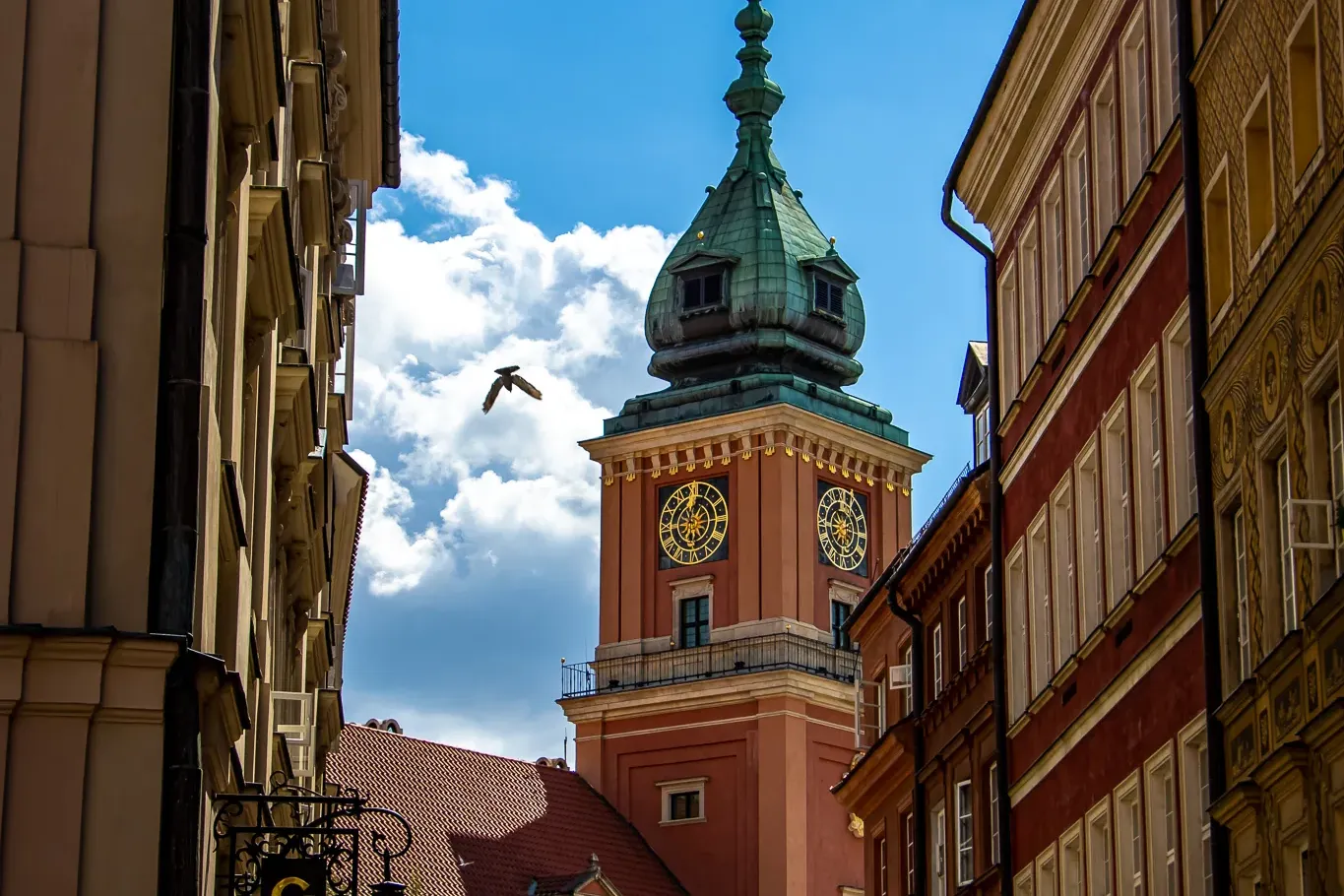 View up a narrow street towards the Royal Castle clock tower in Warsaw with a green dome and golden clock faces, a bird fl...