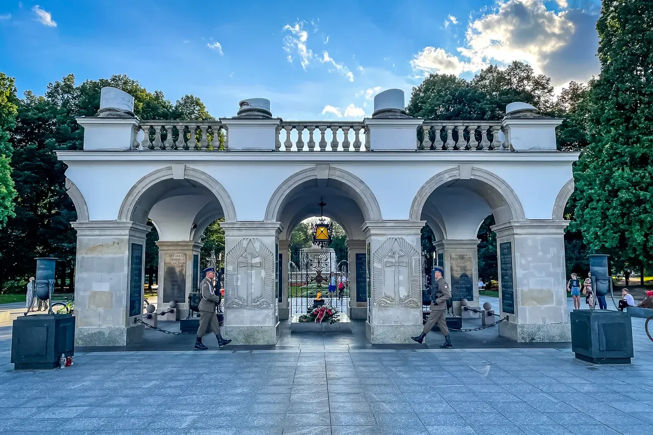 Two uniformed guards march in front of the white stone arcades of the Tomb of the Unknown Soldier in Warsaw under a blue sky.