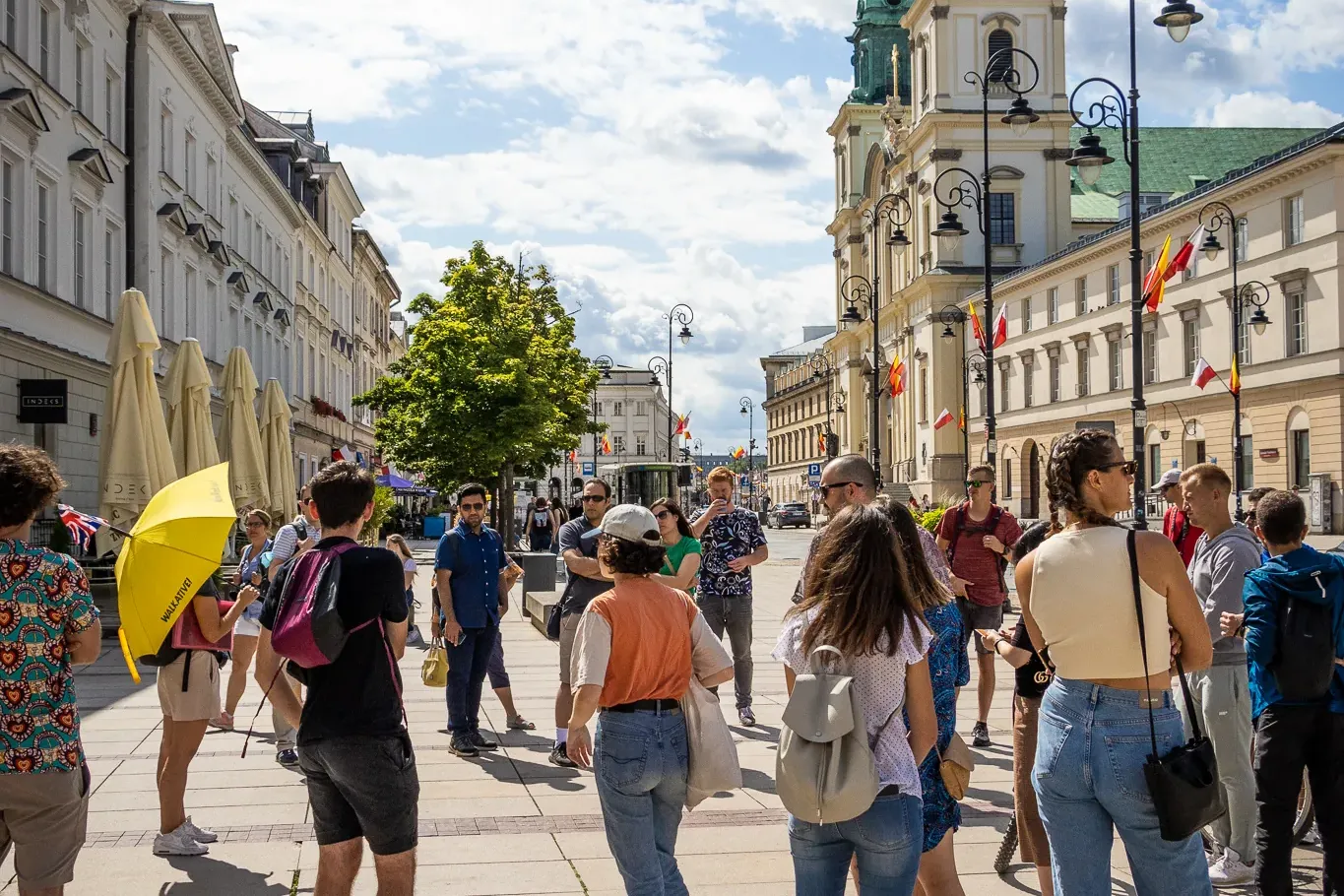 A group of people gathered on a paved street in Warsaw with a historic church and other buildings in the background under ...