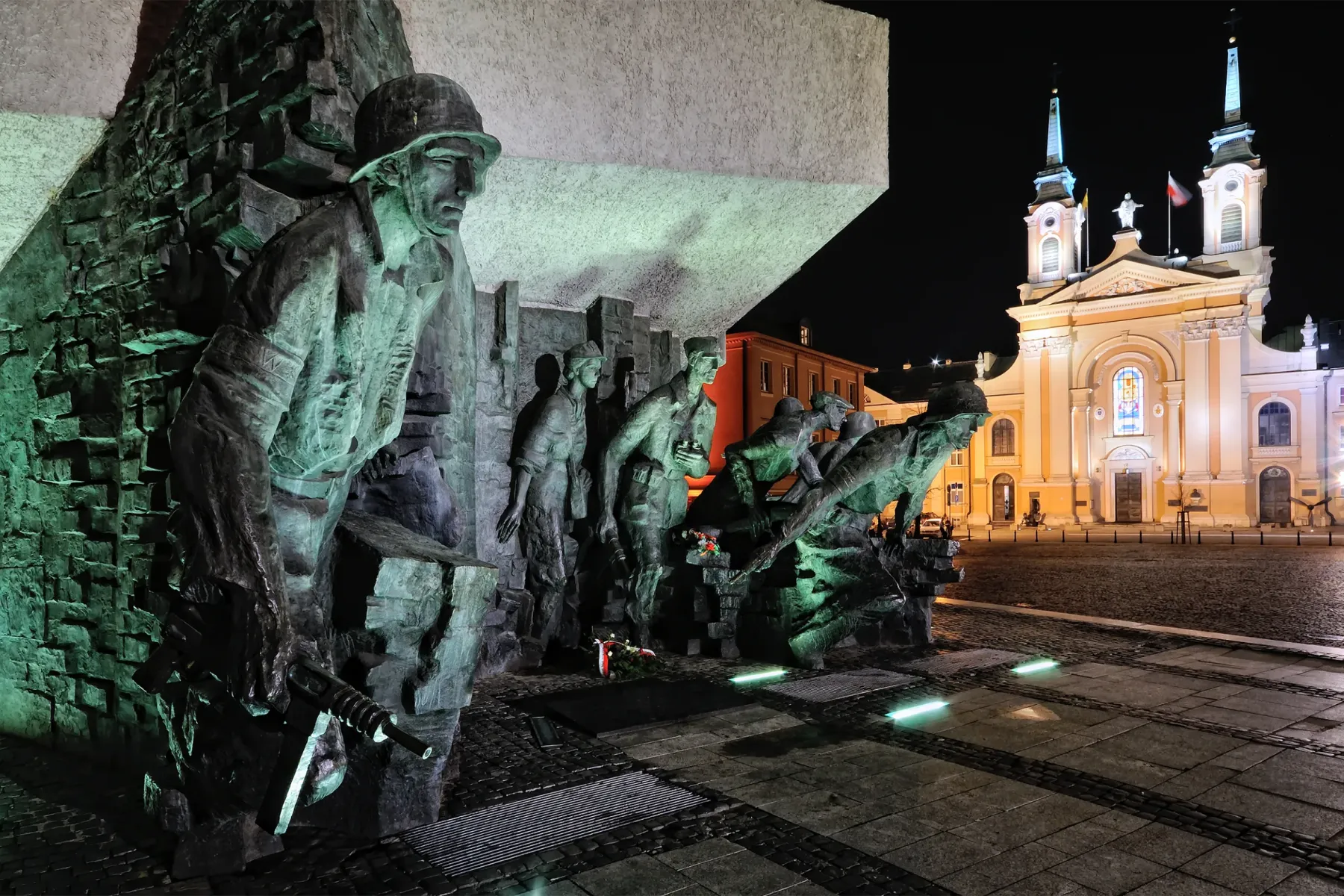 Bronze monument depicting figures of Warsaw Uprising soldiers under green light at night, with a brightly lit church behind.