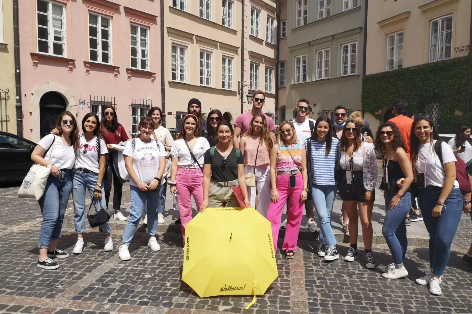 A group of young adults poses on a cobbled street with a yellow Walkative umbrella. Colorful historic buildings in backgro...