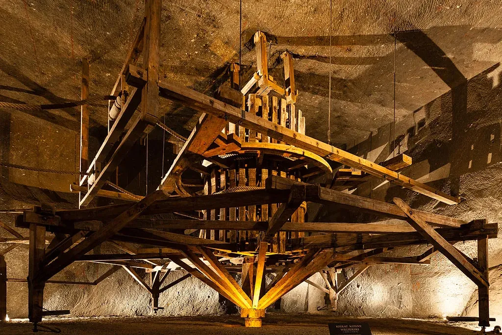 A large, historical wooden horse-powered hoisting machine inside the Wieliczka Salt Mine, with salt rock walls behind it.