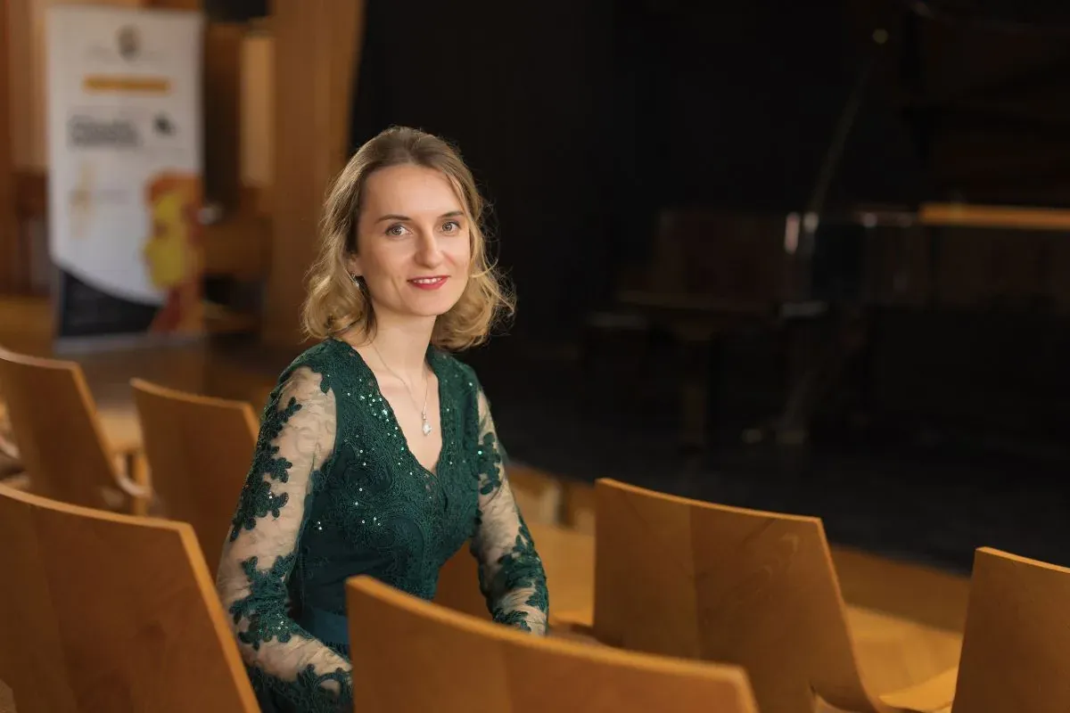 A smiling woman in a dark green dress with lace sleeves sits in a concert hall. Empty wooden chairs and a grand piano are ...