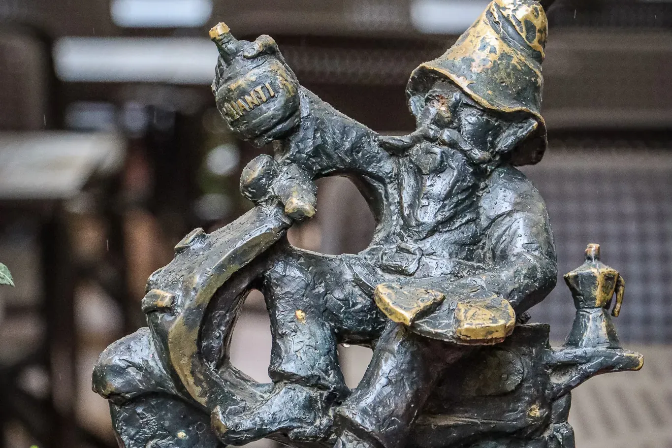 Bronze statue of a Wroclaw dwarf riding a scooter. The dwarf wears a pointy hat and holds a bottle of Chianti. A moka pot ...