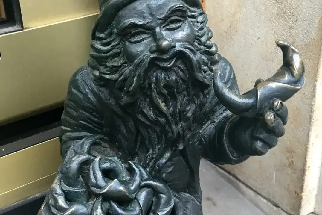 Bronze dwarf statue with a pointed hat and long beard, holding a tray with knots and a horn-shaped object, next to a build...