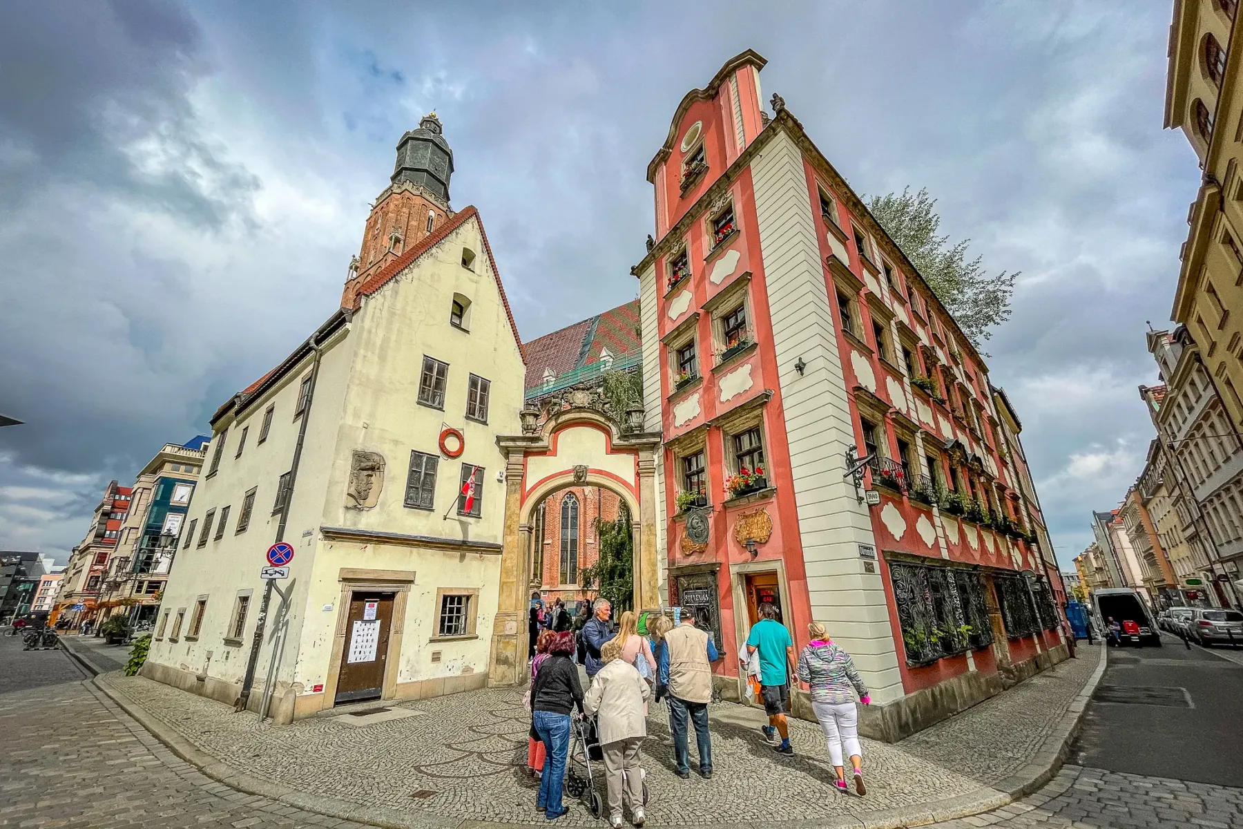 People walk on a cobblestone street in Wroclaw's Old Town, past historic yellow and red buildings, and an archway leading ...