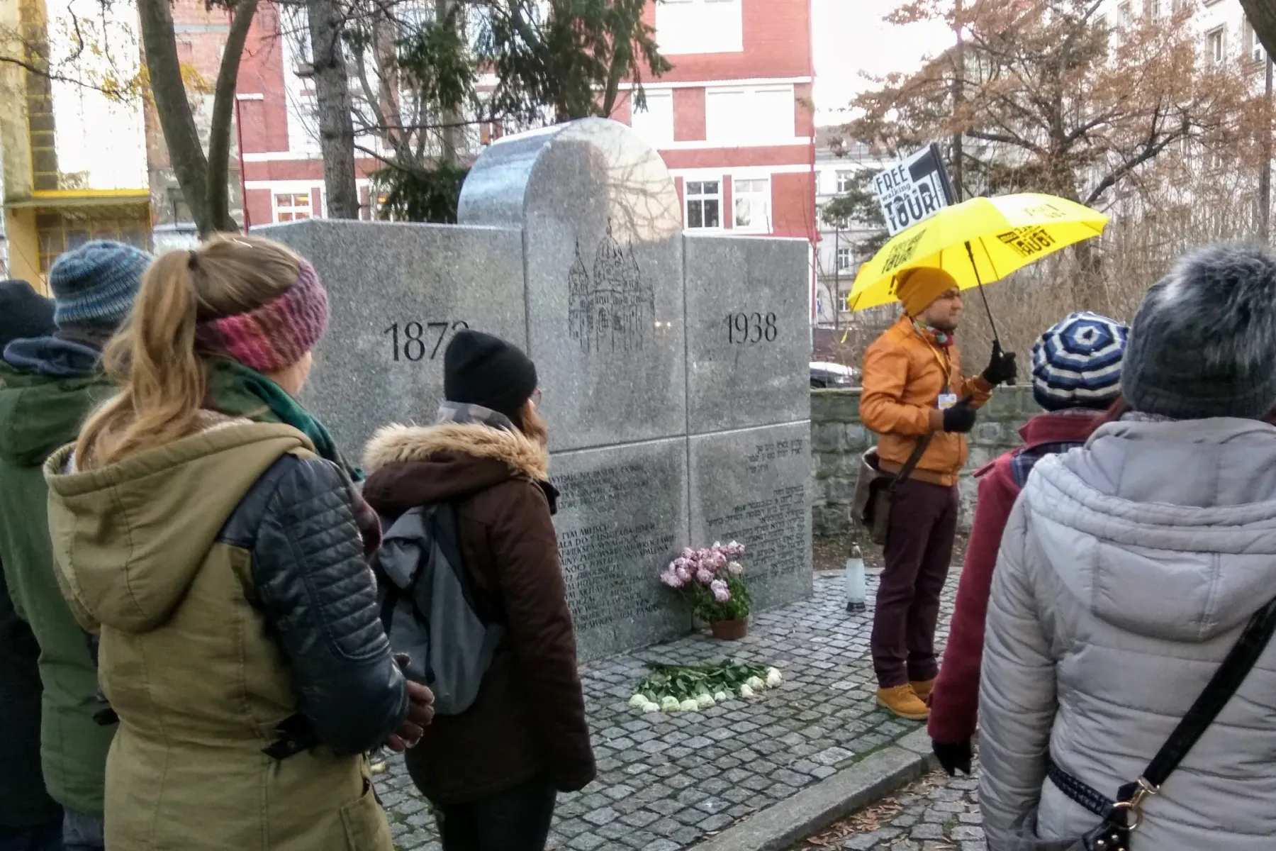 Stone monument commemorating the New Synagogue with dates 1870 and 1938, attended by a tour group with guide.