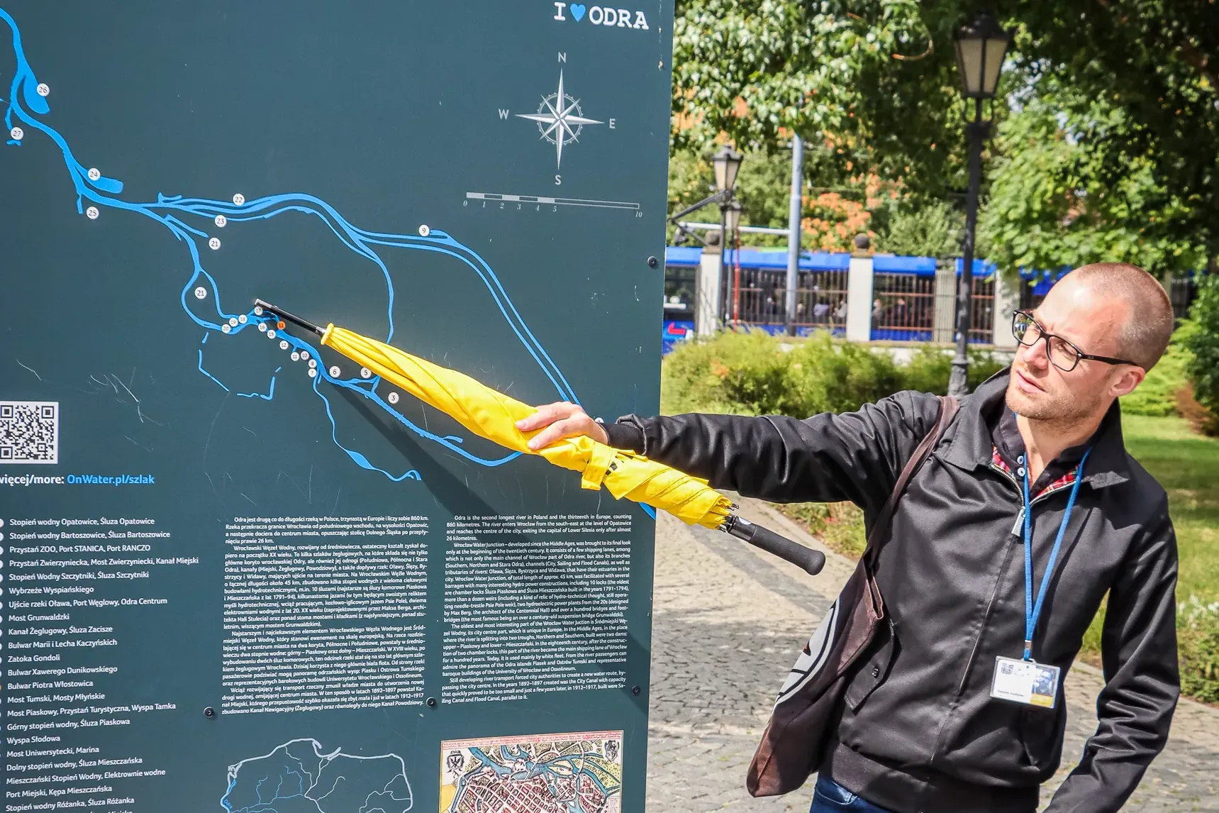 Man pointing with a yellow umbrella at an information board displaying a map of Wrocław's Floodway System on a sunny day.