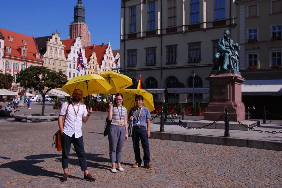 Three people with yellow umbrellas stand on a cobblestone square in front of historic buildings and a statue on a sunny day.