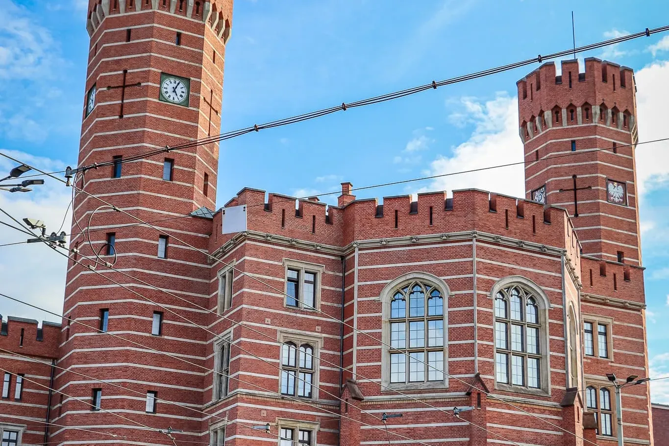 Historic red-brick building with two towers, each featuring a clock face, under a blue sky with white clouds in Wroclaw.