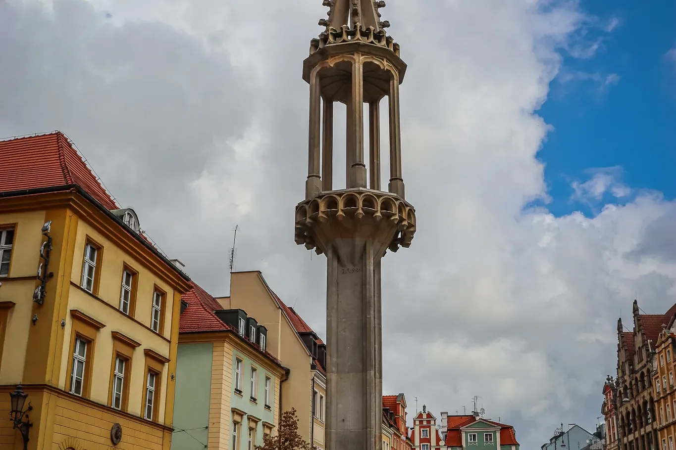 Tall stone pillory with an ornate spire and a figure on top, surrounded by colorful historic buildings in a European city ...