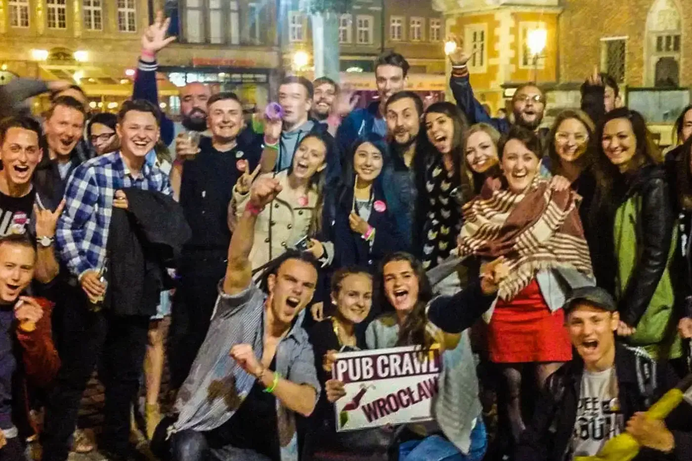 Group of cheerful people at a night pub crawl in Wrocław, with historic buildings in the background. A sign reads "Pub Cra...