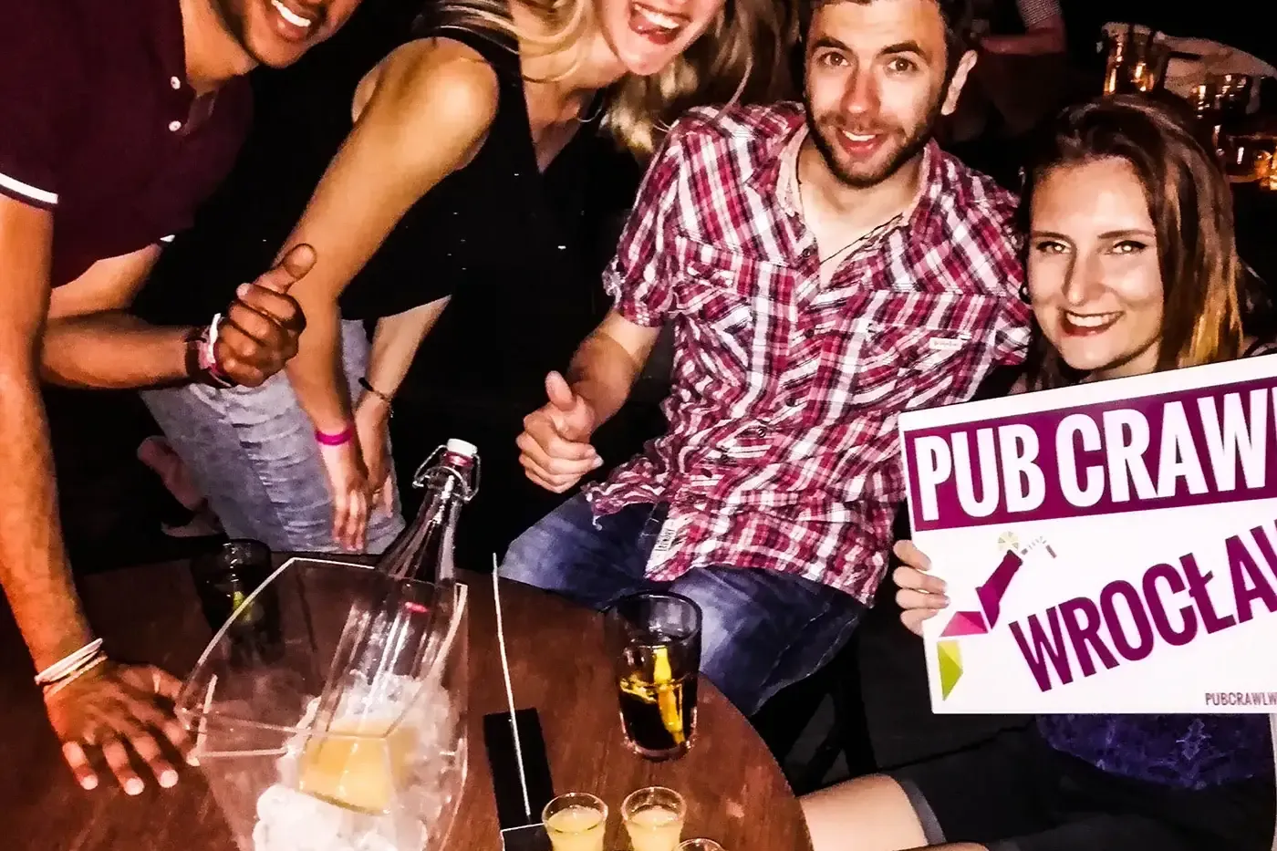 Four smiling young adults at a table with drinks and shots, one holding a 'PUB CRAWL WROCŁAW' sign.