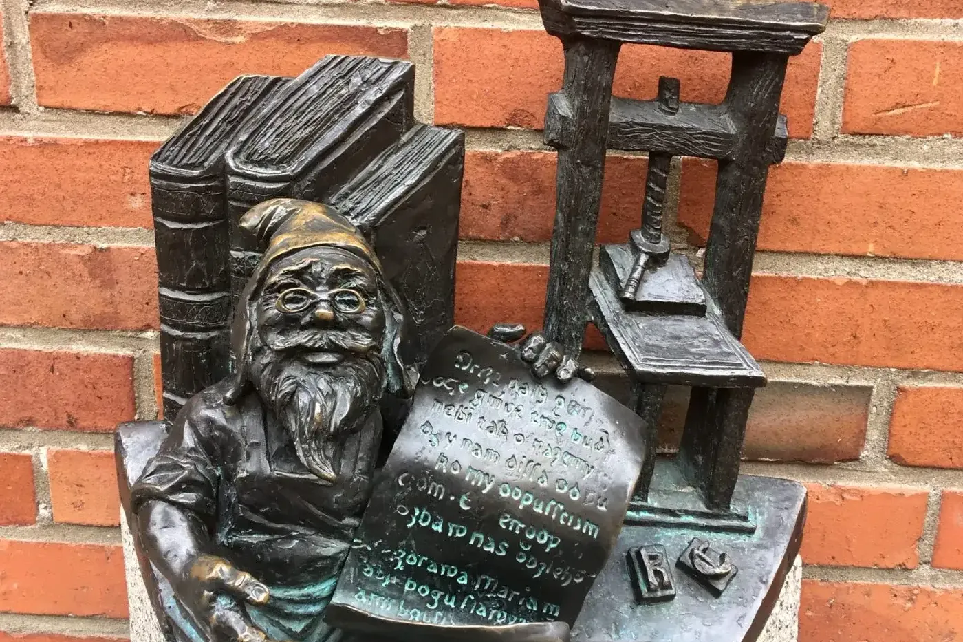Bronze statue of a bespectacled dwarf holding a document, next to books and a printing press, against a red brick wall.