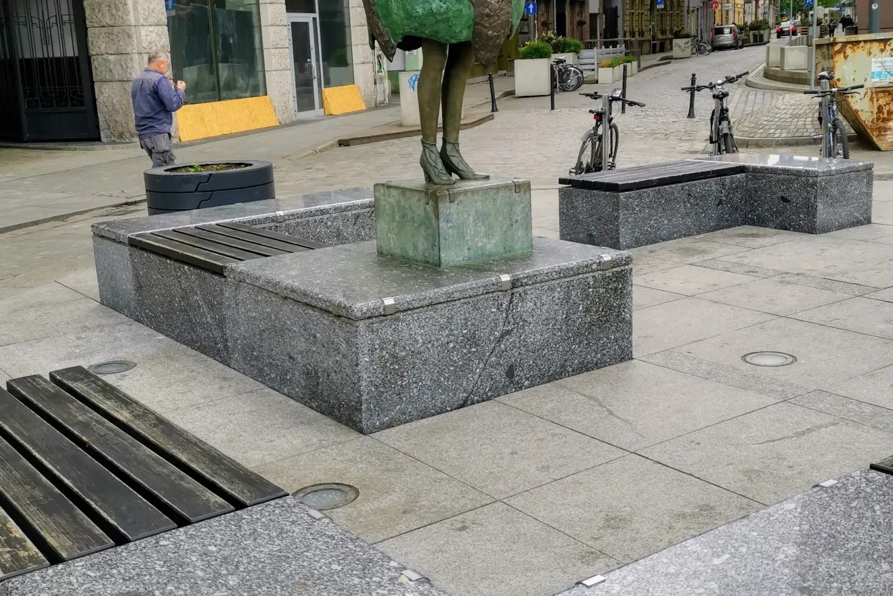 Bronze statue of a woman with a globe as a skirt, granite benches, and a metal plaque with religious symbols on a city str...