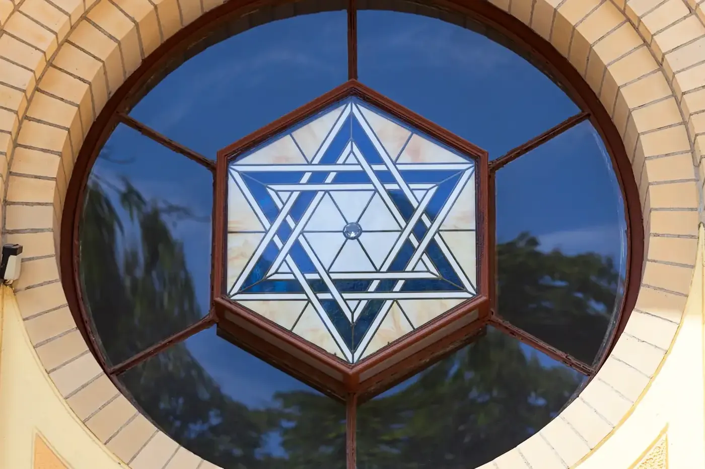 Round stained glass window with a blue and white Star of David design, set in a yellow brick arch above a brown wooden frame.