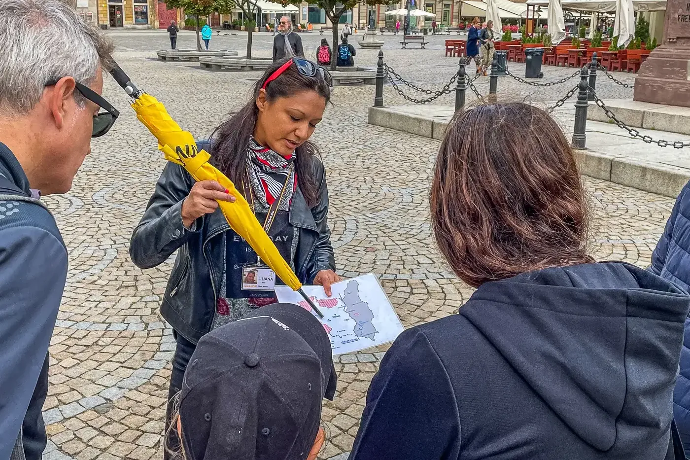 Female tour guide with yellow umbrella and map explains to three tourists on a cobblestone square. Colorful historic build...