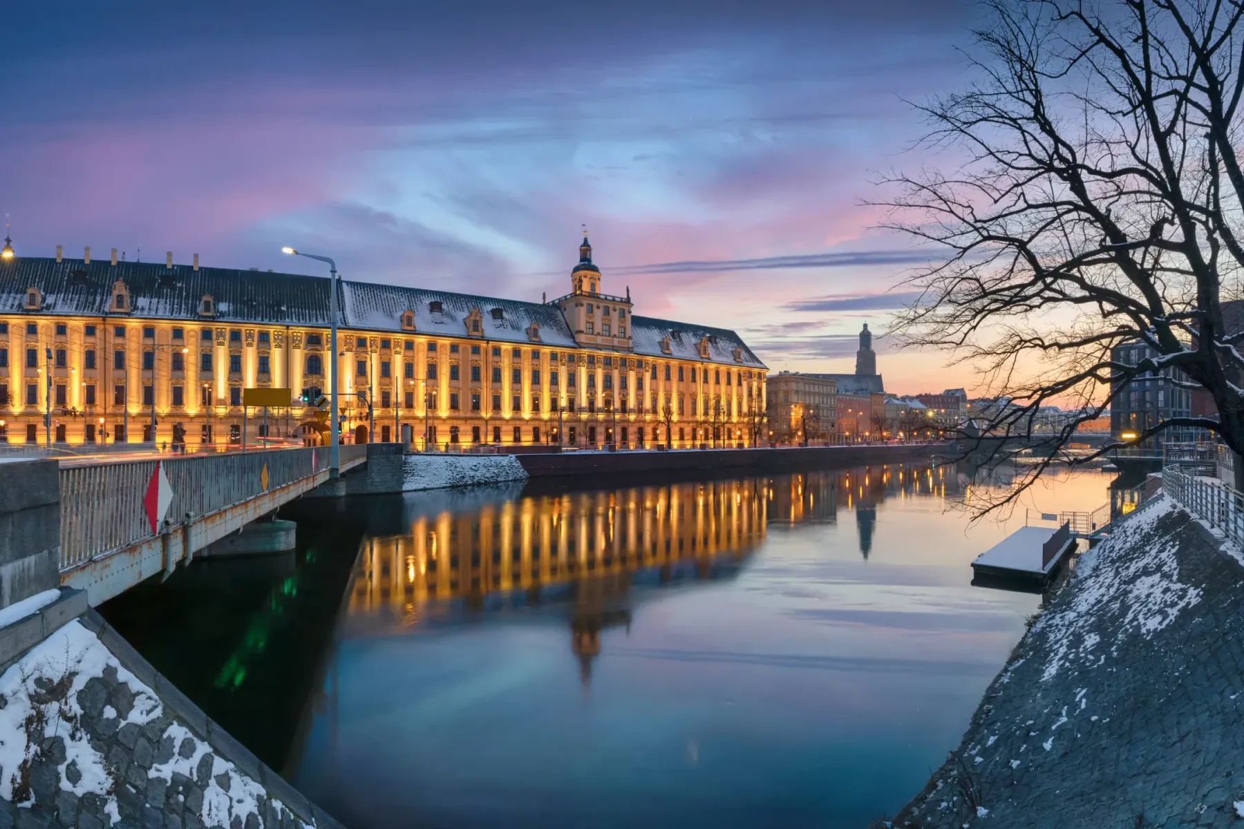 Illuminated University of Wrocław building and bridge over the Odra River at dusk, with snow on banks.