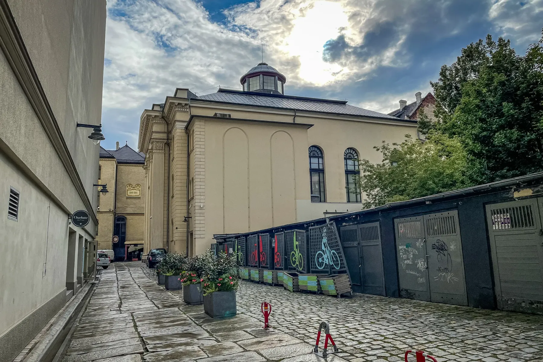 Cobblestone alleyway leading to the White Stork Synagogue in Wroclaw under a cloudy sky. Potted plants and bicycle racks a...