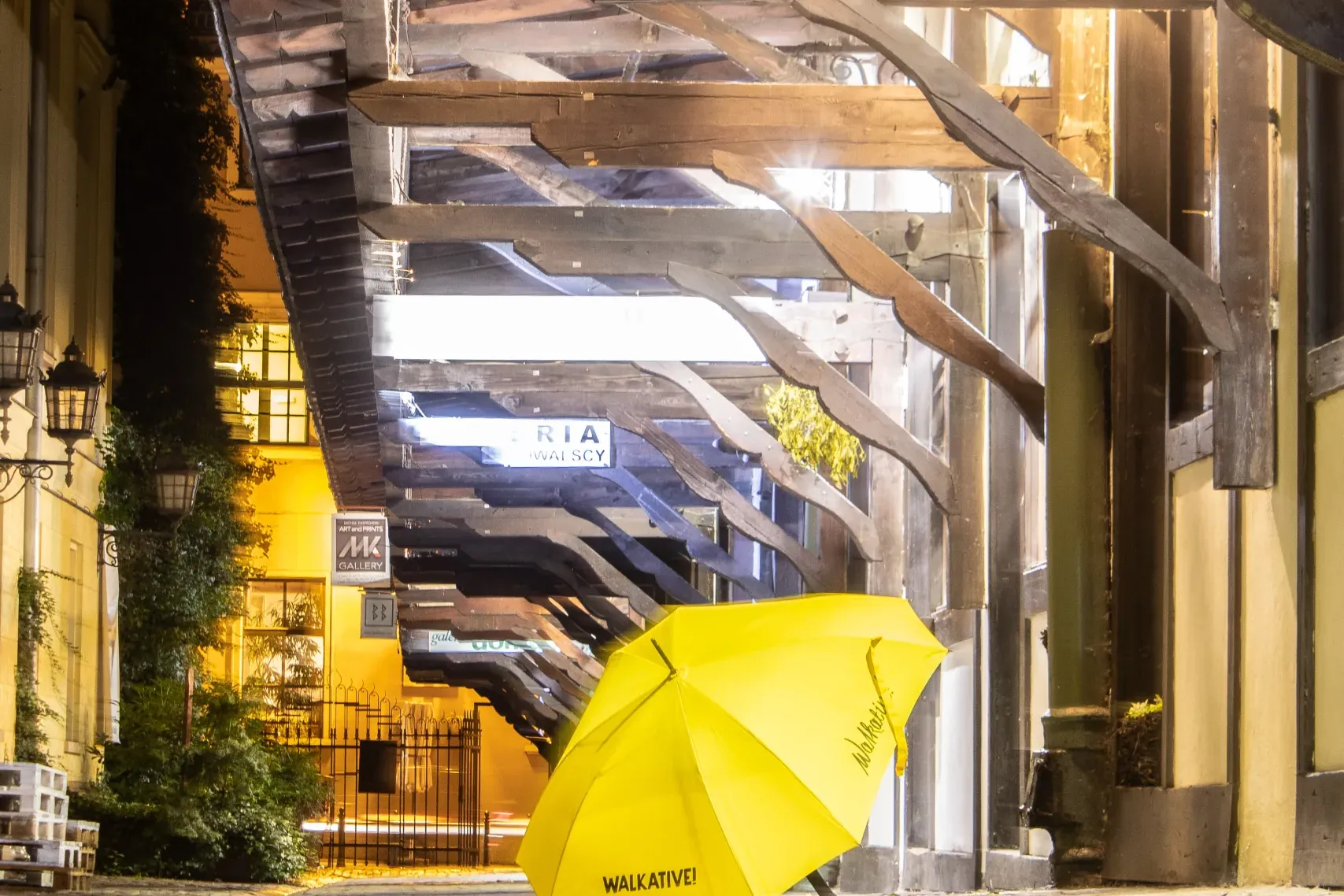 Yellow Walkative! umbrella on a cobblestone street at night, beneath a timber-framed passage illuminated by a bright light.