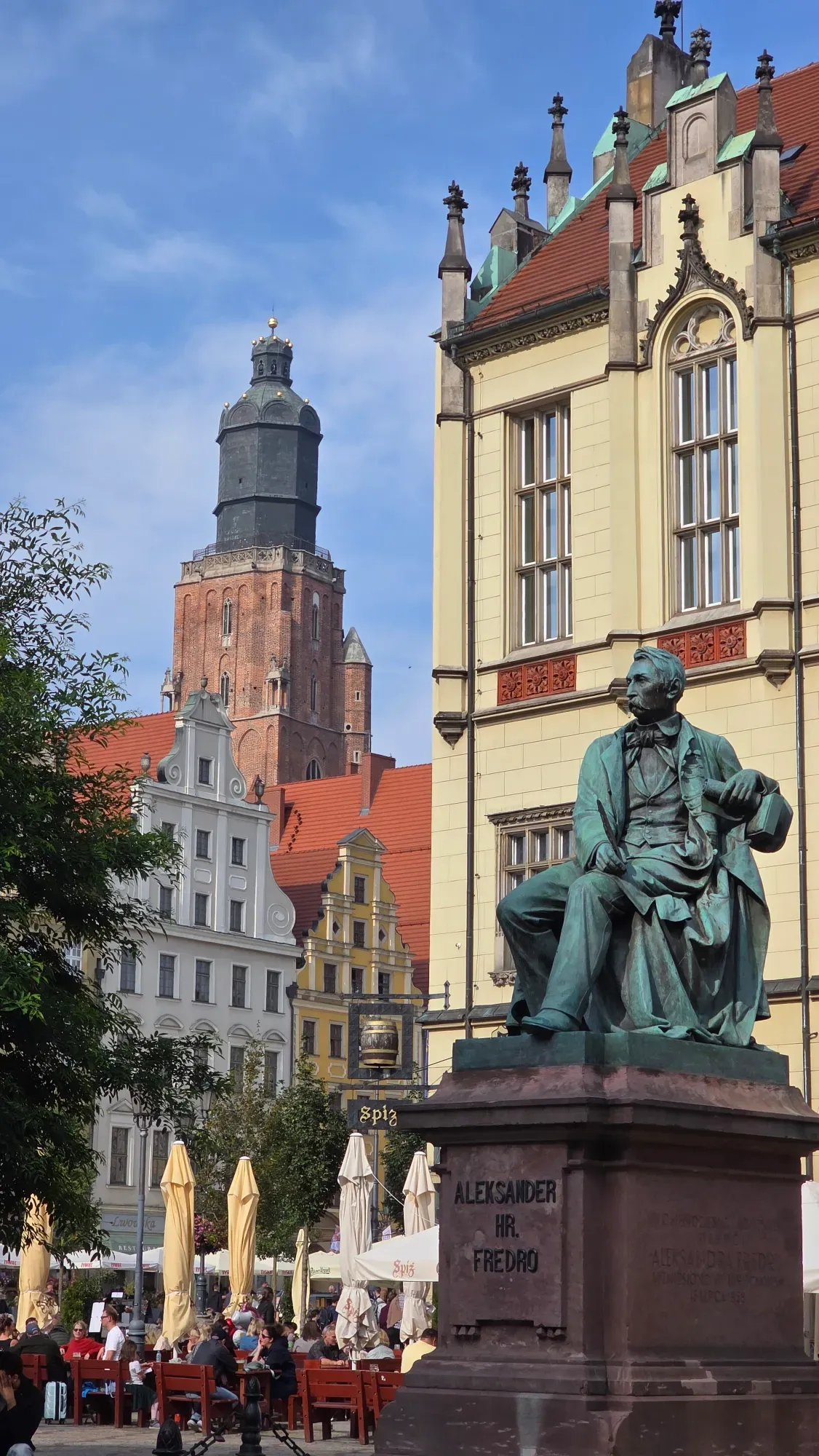 A bronze statue of Aleksander Fredro in Wrocław's Market Square, with historic buildings and a church tower in the backgro...