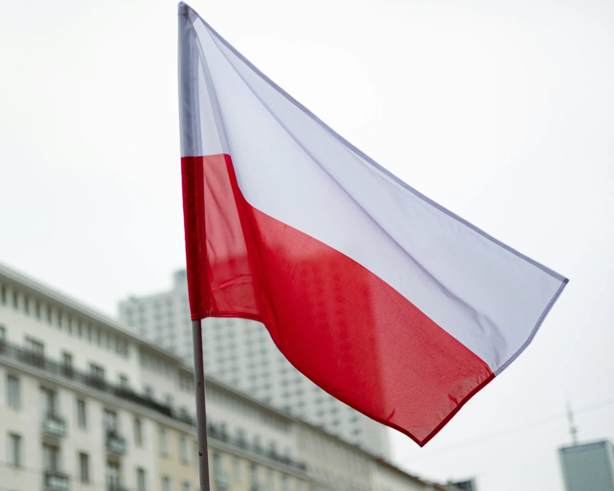 Polish national flag with white and red horizontal stripes waving against city buildings.