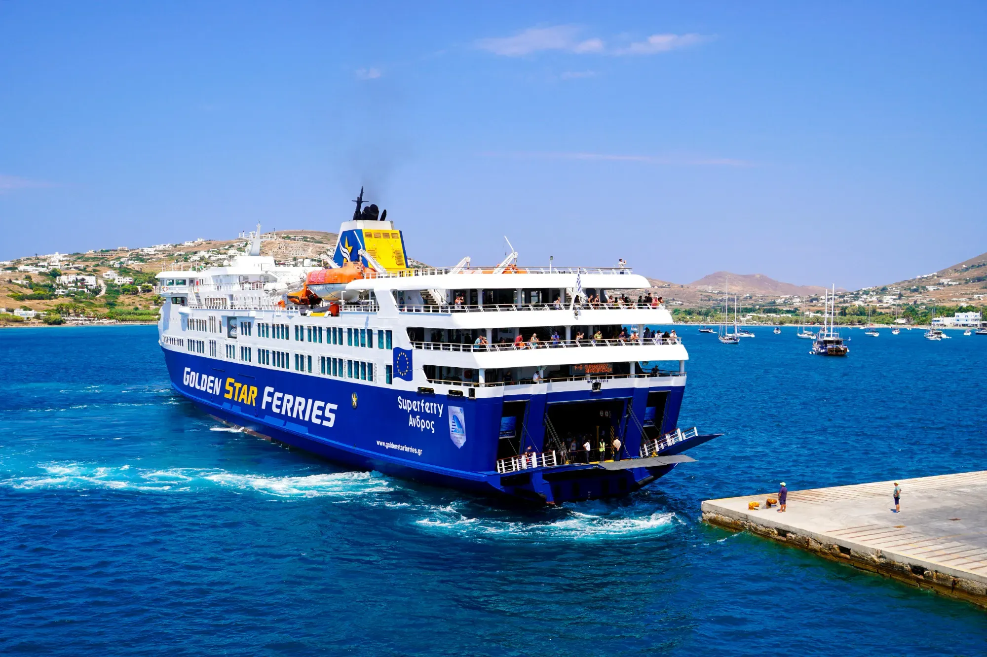 Large blue and white ferry departing a concrete pier, creating a wake. Passengers on deck. Distant coast with buildings under